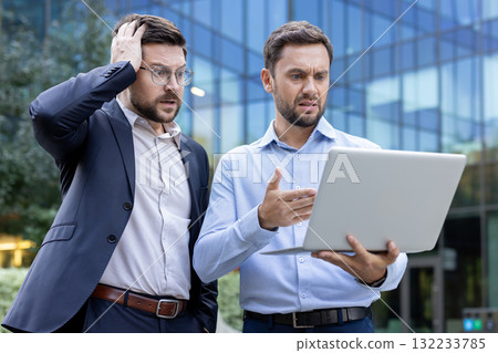 Two stressed businessmen in business casual attire reacting to shocking bad news on a laptop screen, expressing confusion and frustration outdoors in front of a modern office building Two stressed businessmen in business casual attire reacting to shocking bad news on a laptop screen, expressing confusion and frustration outdoors in front of a modern office building 132233785
