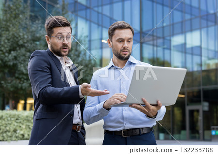 Two concerned businessmen standing outdoors in front of a modern office building, reacting with surprise and frustration to a serious issue displayed on a laptop computer 132233788