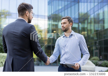 Two professional businessmen shaking hands outside a modern office building, symbolizing agreement, successful negotiation, and a new partnership forming between colleagues during a business meeting 132233800