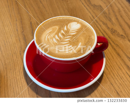 Cup of cappuccino with latte art in red cup on wooden table. Warmth, aroma, and comfort in cozy cafe atmosphere. 132233830