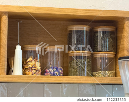 Glass jars with dried herbs and fruits on wooden shelf. Natural ingredients, tea blends and cozy cafe interior atmosphere. 132233833