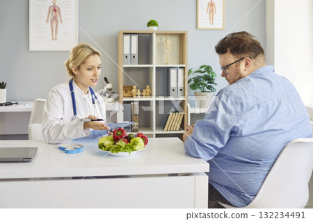 Nutritionist consulting overweight patient with vegetables, tape measure in clinic 132234491