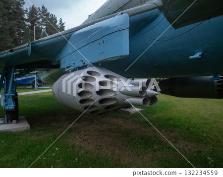rocket cluster mounted under wing near tree line, close view of multiple barrels and launch tubes with camouflage paint and wet grass, evocative of tactical hardware and cold, heavy rocket cluster mounted under wing near tree line, close view of multiple barrels and launch tubes with camouflage paint and wet grass, evocative of tactical hardware and cold, heavy 132234559