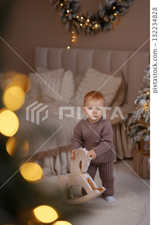 Toddler boy holding wooden rocking horse near Christmas tree in cozy beige bedroom 132234828