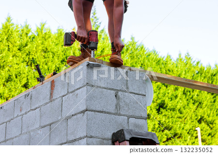 Construction worker drills into wooden beams while standing on cinder block wall Construction worker drills into wooden beams while standing on cinder block wall 132235052