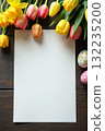 Colorful tulips and painted Easter eggs surround empty white sheet on dark wood table, perfect for spring or holiday-themed message or design Space for text 132235200