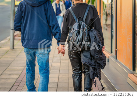 Young couple walks hand in hand on sidewalk, dressed in casual streetwear and leather. Concept of love, relationship, urban romance, casual urban lifestyle Young couple walks hand in hand on sidewalk, dressed in casual streetwear and leather. Concept of love, relationship, urban romance, casual urban lifestyle 132235362