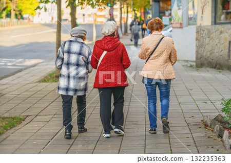 Senior women. Three elderly women walking together on city sidewalk in warm autumn clothes, back view. Concept of senior friendship and everyday urban life Senior women. Three elderly women walking together on city sidewalk in warm autumn clothes, back view. Concept of senior friendship and everyday urban life 132235363