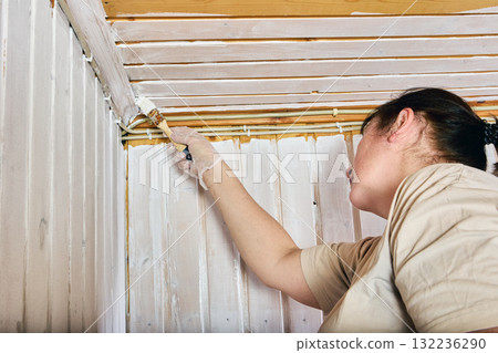 Coating edge, female painter applying white layer along meeting line of vertical and horizontal wood paneling to achieve consistent interior finish. Coating edge, female painter applying white layer along meeting line of vertical and horizontal wood paneling to achieve consistent interior finish. 132236290