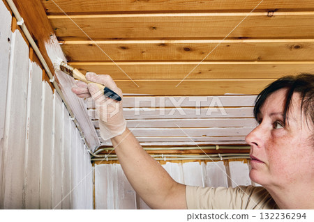 Painting corner, woman carefully brushing white paint on junction between wall paneling and upper timber boards to complete uniform surface coating. Painting corner, woman carefully brushing white paint on junction between wall paneling and upper timber boards to complete uniform surface coating. 132236294
