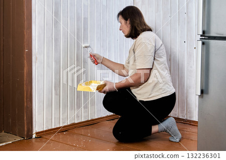 Walls seem partially coated near completion with white paint, painter works from low position using small roller, holding yellow tray close to tongue and groove panels. Walls seem partially coated near completion with white paint, painter works from low position using small roller, holding yellow tray close to tongue and groove panels. 132236301