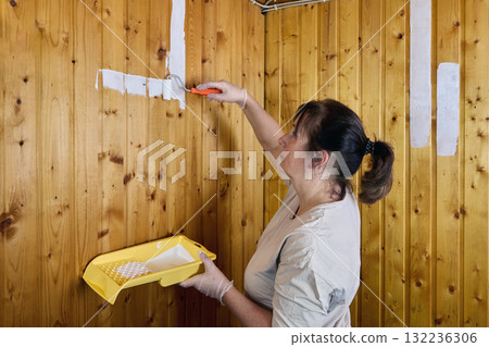 Painting with roller creates wide white stripe across vertical wooden panels, female painter holds yellow tray near wall while applying first layer of paint. 132236306