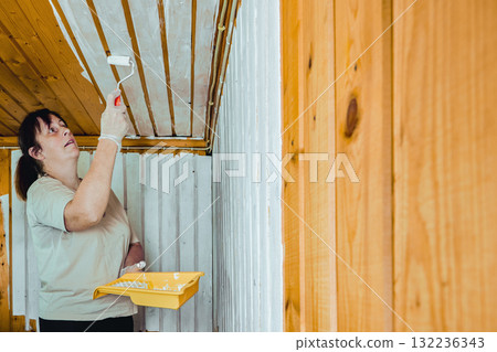 Painting paneling, woman using roller to coat upper wooden wall section with white paint continuing process of covering entire interior surface. Painting paneling, woman using roller to coat upper wooden wall section with white paint continuing process of covering entire interior surface. 132236343