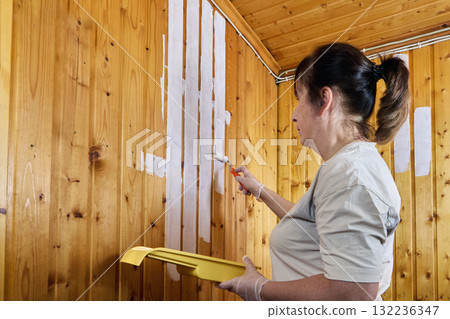 Woman painter applies white paint to wall covered with paneling holding paint tray in left hand and mini roller in right. Woman painter applies white paint to wall covered with paneling holding paint tray in left hand and mini roller in right. 132236347