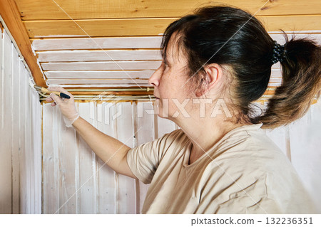 Decorating job, female worker covering timber cladding with white paint brush strokes transforming natural boards into bright finish. 132236351