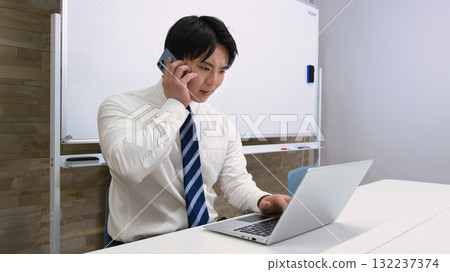 A young businessman talking on a smartphone while looking at a computer in the office A young businessman talking on a smartphone while looking at a computer in the office 132237374