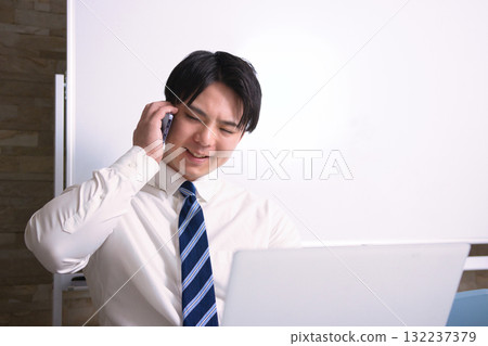 A young businessman talking on a smartphone while looking at a computer in the office A young businessman talking on a smartphone while looking at a computer in the office 132237379