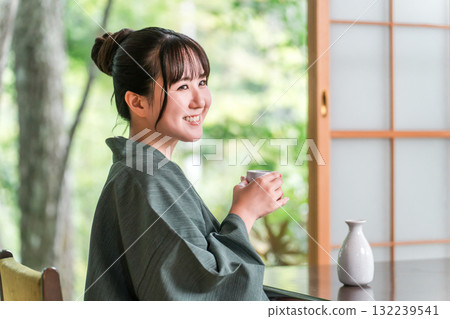 A young Asian woman in a yukata haori relaxing on the veranda of a guest room in a ryokan, drinking sake A young Asian woman in a yukata haori relaxing on the veranda of a guest room in a ryokan, drinking sake 132239541