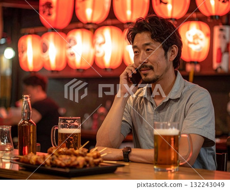 一名男子在一家掛著紅燈籠的熱門居酒屋裡喝啤酒、打電話 132243049