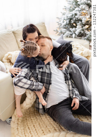 A loving family shares a tender moment with their dog in front of a Christmas tree. 132247381