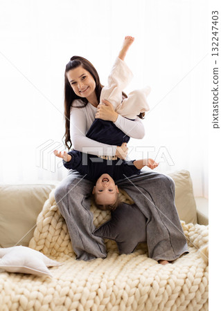 A mother holds her child upside down on a cozy couch, both smiling and enjoying a playful moment together. 132247403