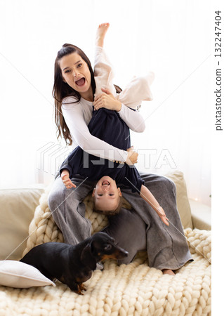 A mother joyfully plays with her child, holding them upside down on a cozy couch, with a dog nearby. 132247404