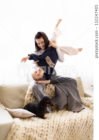 A mother playfully holds her child upside down on a cozy couch, with a dachshund dog nearby. 132247405