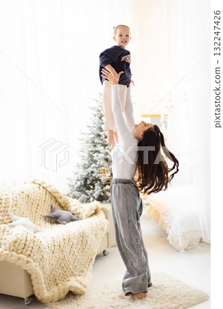 A mother joyfully lifts her child up in the air, creating a moment of happiness and connection in a bright, cozy room. A mother joyfully lifts her child up in the air, creating a moment of happiness and connection in a bright, cozy room. 132247426