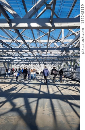 Pedestrians walk across a modern steel-framed bridge in Saint Petersburg on a bright day, creating strong geometric shadows on the ground. Pedestrians walk across a modern steel-framed bridge in Saint Petersburg on a bright day, creating strong geometric shadows on the ground. 132248119