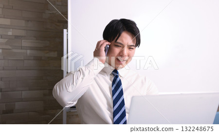 一位年輕的商人在辦公室裡一邊看著電腦一邊用智慧型手機聊天 132248675