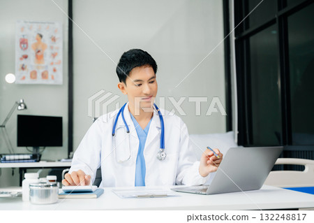 Confident young  male doctor in white medical uniform sit at desk working on computer. Smiling use laptop write in medical journal 132248817