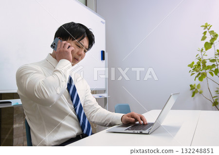 一位年輕的商人在辦公室裡一邊看著電腦一邊用智慧型手機聊天 132248851