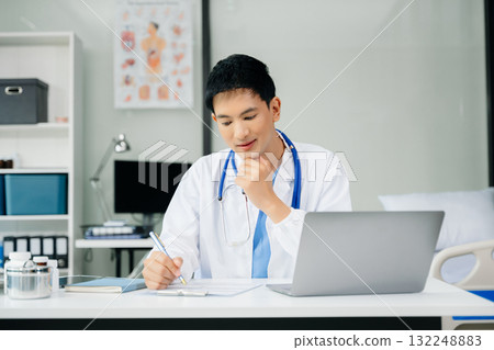 Confident young male doctor in white medical uniform sit at desk working on computer. Smiling use laptop write in medical journal Confident young male doctor in white medical uniform sit at desk working on computer. Smiling use laptop write in medical journal 132248883