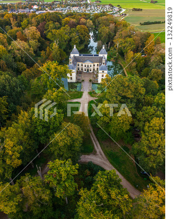 Aerial view of historic castle symmetrical twin towers and central hall framed by forest, moats, and tree lined path, with open fields and residential zone beyond under warm sunrise sky. Aerial view of historic castle symmetrical twin towers and central hall framed by forest, moats, and tree lined path, with open fields and residential zone beyond under warm sunrise sky. 132249819