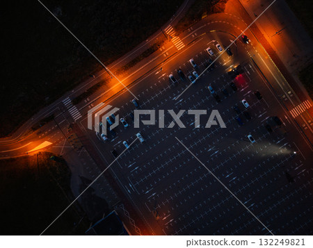 Aerial nighttime view of empty parking lot warm orange streetlights illuminate marked lanes and scattered cars, highlighting symmetry 132249821