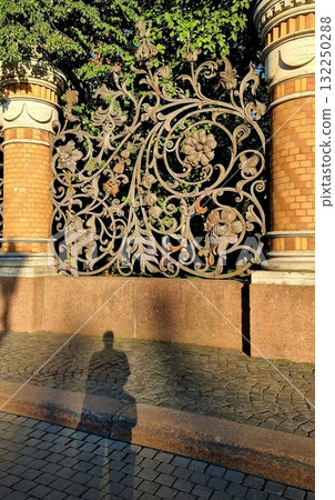 An ornate cast-iron fence panel with floral motifs and brick columns in Saint Petersburg, with a long shadow of a person on the sidewalk. An ornate cast-iron fence panel with floral motifs and brick columns in Saint Petersburg, with a long shadow of a person on the sidewalk. 132250288