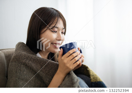 Young woman enjoying a hot drink under a blanket by the window Young woman enjoying a hot drink under a blanket by the window 132252394