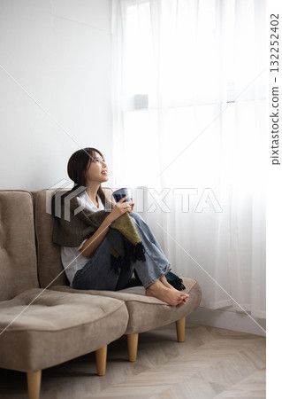 Young woman enjoying a hot drink under a blanket by the window Young woman enjoying a hot drink under a blanket by the window 132252402