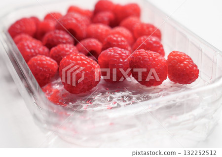 Fresh raspberries in plastic transparent supermarket packaging on a white table surface Fresh raspberries in plastic transparent supermarket packaging on a white table surface 132252512