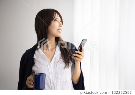 一位微笑的女士在窗邊拿著杯子，玩著智慧型手機 132252592