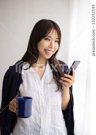 一位微笑的女士在窗邊拿著杯子，玩著智慧型手機 132252593