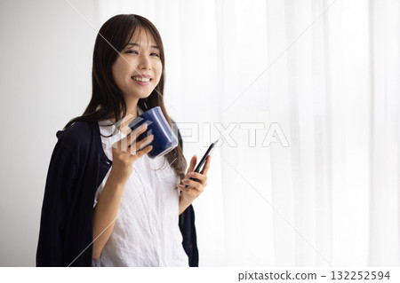 一位微笑的女士在窗邊拿著杯子，玩著智慧型手機 132252594