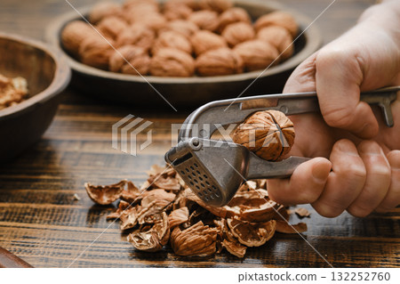 Hand using a nutcracker to shell walnuts on a wooden table with more walnuts in the background Hand using a nutcracker to shell walnuts on a wooden table with more walnuts in the background 132252760