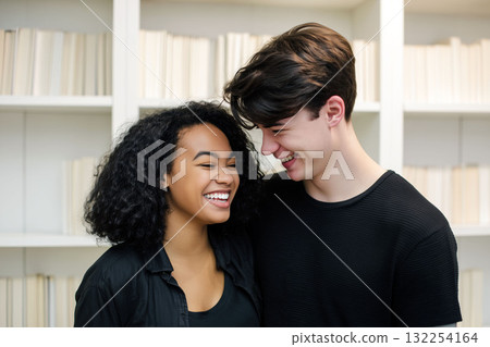 Young couple laughing together in front of a bookshelf, sharing a joyful moment in a cozy indoor setting 132254164