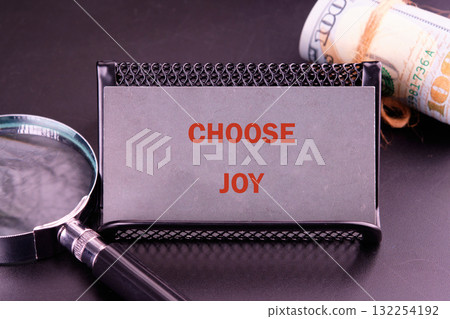 A sleek business card holder displays the message Choose Joy, surrounded by a magnifying glass and rolled money, blending motivation with finance in a modern workspace setting A sleek business card holder displays the message Choose Joy, surrounded by a magnifying glass and rolled money, blending motivation with finance in a modern workspace setting 132254192