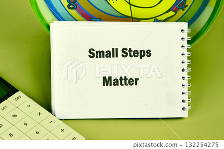 A notepad displaying the phrase Small Steps Matter sits beside a calculator and a colorful clock. This scene represents the importance of gradual progress in achieving goals and success A notepad displaying the phrase Small Steps Matter sits beside a calculator and a colorful clock. This scene represents the importance of gradual progress in achieving goals and success 132254275
