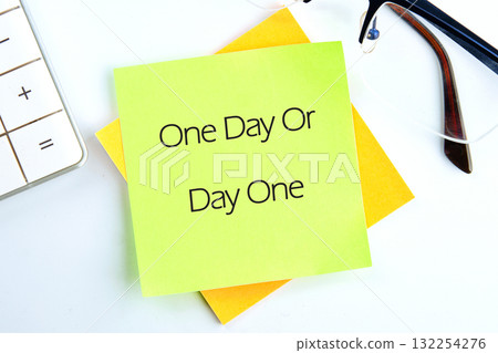 A vibrant sticky note displays the phrase One Day Or Day One, encouraging decision-making and action, placed on a tidy desk alongside glasses and a calculator in daylight A vibrant sticky note displays the phrase One Day Or Day One, encouraging decision-making and action, placed on a tidy desk alongside glasses and a calculator in daylight 132254276