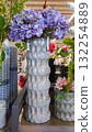 Decorative vase with textured surface, filled with vibrant purple hydrangeas, surrounded by various floral arrangements in a bright, inviting retail environment showcasing home decor 132254889