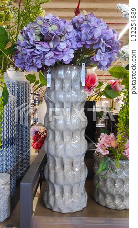 Decorative vase with textured surface, filled with vibrant purple hydrangeas, surrounded by various floral arrangements in a bright, inviting retail environment showcasing home decor Decorative vase with textured surface, filled with vibrant purple hydrangeas, surrounded by various floral arrangements in a bright, inviting retail environment showcasing home decor 132254889