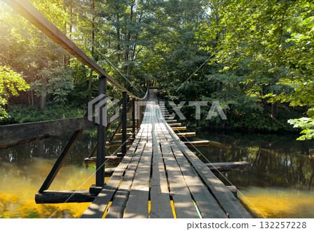 Bridge stretches over sunny rural river. Wooden suspended footbridge with rope sides reflects in Bridge stretches over sunny rural river. Wooden suspended footbridge with rope sides reflects in 132257228